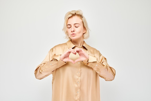 Mulher de camisa bege fazendo forma de coração com as mãos em fundo branco