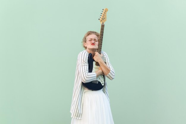 Mulher de camisa branca e saia blusa com óculos em pé com os olhos fechados e abraçando a guitarra