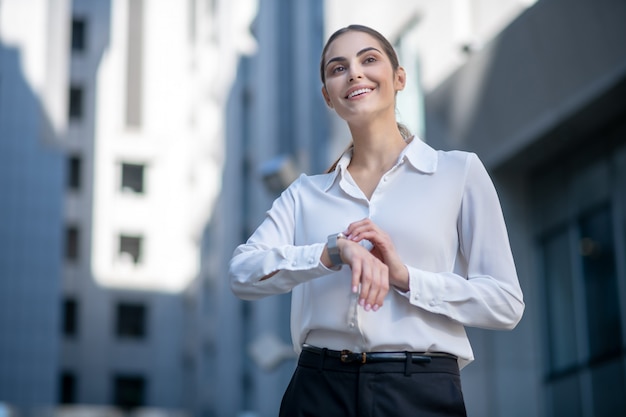 Mulher de camisa branca tocando um smartwatch e parecendo satisfeita