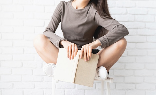 Foto mulher de camisa marrom sentada na cadeira com as pernas cruzadas segurando um livro aberto