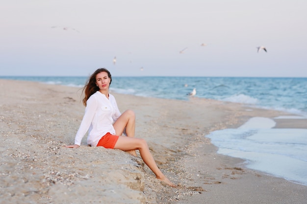 Mulher de camiseta branca e short vermelho está sentada perto do mar em uma praia no verão.