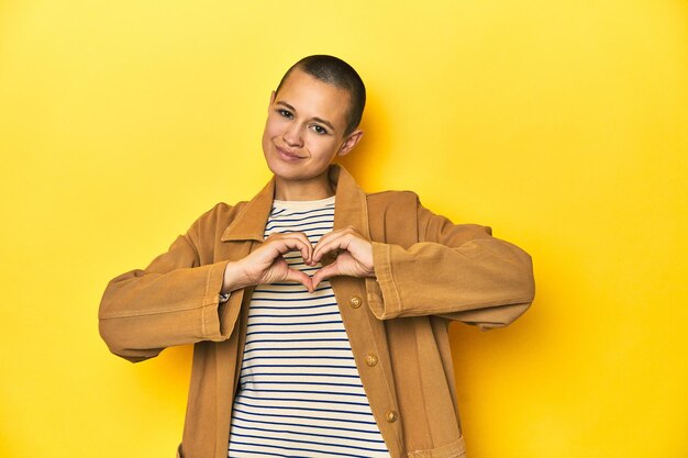 Mulher de camiseta listrada e camisa jeans com fundo amarelo sorrindo e mostrando uma forma de coração com as mãos