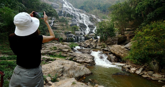 Mulher de camiseta preta, tirando fotos da bela cachoeira pelo smartphone.