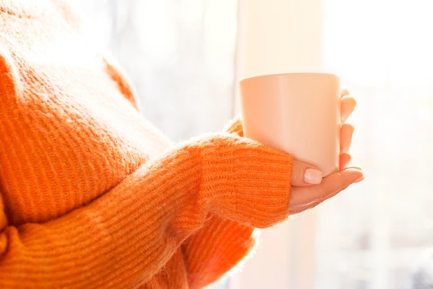 Mulher de camisola laranja segurando uma chávena de chá ou café perto da janela em casa