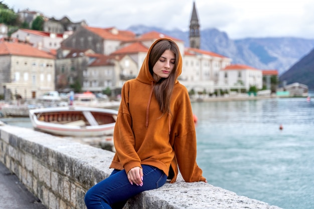 Mulher de capuz laranja fica à beira-mar na cidade Perast em segundo plano no Montenegro