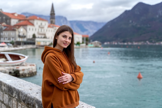 Mulher de capuz laranja fica à beira-mar na cidade Perast em segundo plano no Montenegro