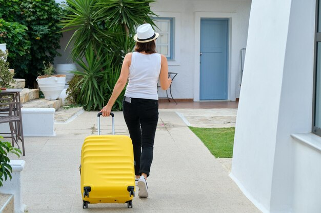 Mulher de chapéu com mala andando ao ar livre pelo território do mar pitoresco hotel spa resort, vista traseira. Viajar, férias, lazer, fim de semana, pessoas