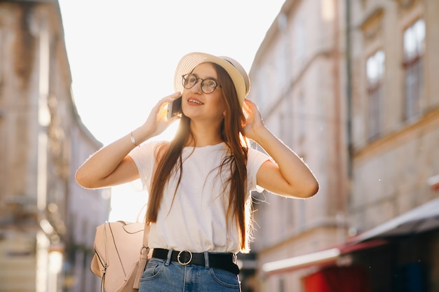 Mulher de chapéu e óculos fica na rua da cidade, falando no celular, sorrindo, rindo.