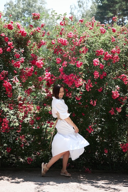 Mulher de chapéu e vestido branco com espartilho caminha no jardim contra o fundo de arbustos rosa Linda garota romântica
