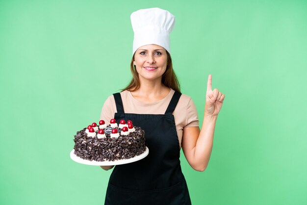 Mulher de chef de pastelaria de meia-idade segurando um bolo grande sobre fundo isolado apontando para cima uma ótima ideia