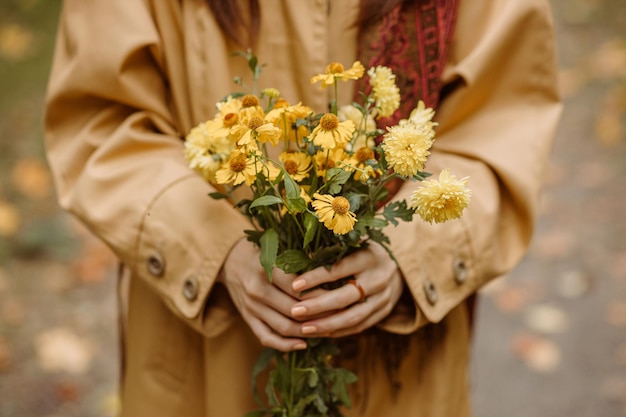 Mulher de colheita na trincheira de outono com buquê de flores frescas amarelas nas mãos em pé no parque