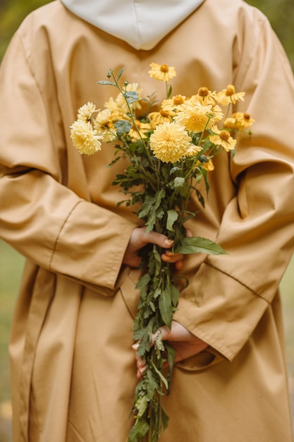 Mulher de colheita na trincheira de outono segurando o buquê de flores amarelas nas costas na natureza