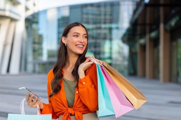 Mulher de compras com sacos de compras na cidade segurando um celular chamando um táxi enquanto está de pé ao ar livre