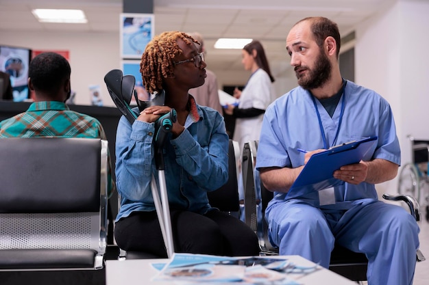 Mulher de consultoria médica com muletas no saguão da recepção do hospital, falando sobre diagnóstico e medicina na sala de espera. Diversas pessoas discutindo na consulta de check-up.