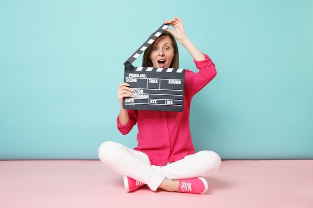 Mulher de corpo inteiro em calça branca de camisa rosa sentada no chão com claquete fazendo filme