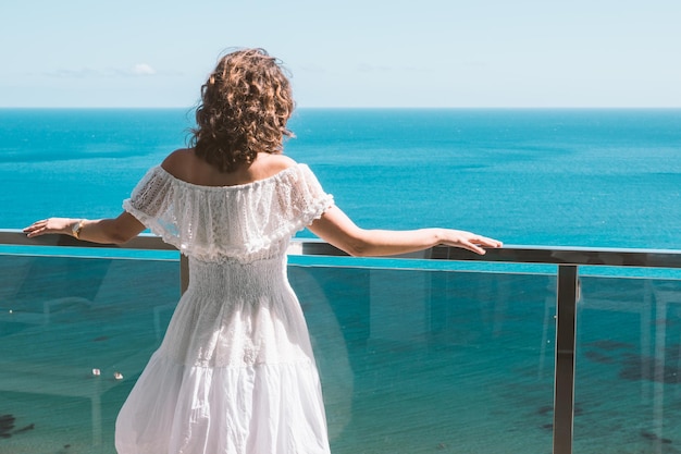 Mulher de costas em um vestido branco de verão com os braços estendidos na grade de uma varanda olhando para o mar azul