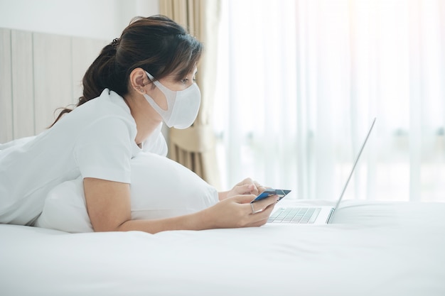 Mulher de doença com máscara facial segurando o cartão de crédito e usando o computador portátil para compras on-line na cama na manhã em casa.