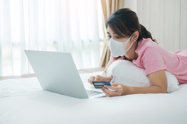 Mulher de doença com máscara facial segurando o cartão de crédito e usando o computador portátil para compras on-line na cama na manhã em casa.