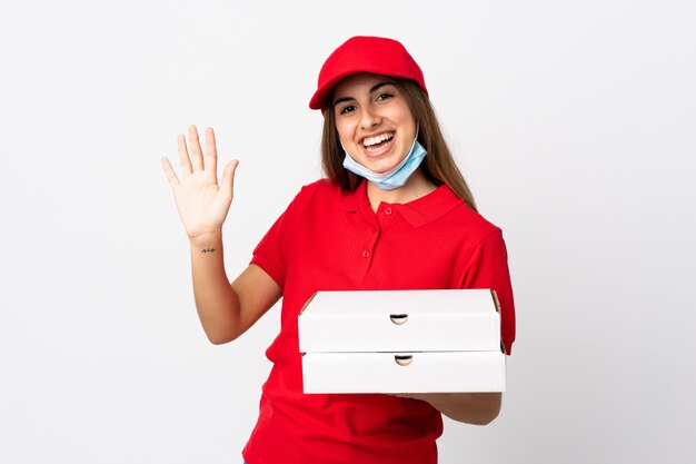 Mulher de entrega de pizza segurando uma pizza e protegendo do coronavírus com uma máscara sobre parede branca isolada, saudando com a mão com expressão feliz