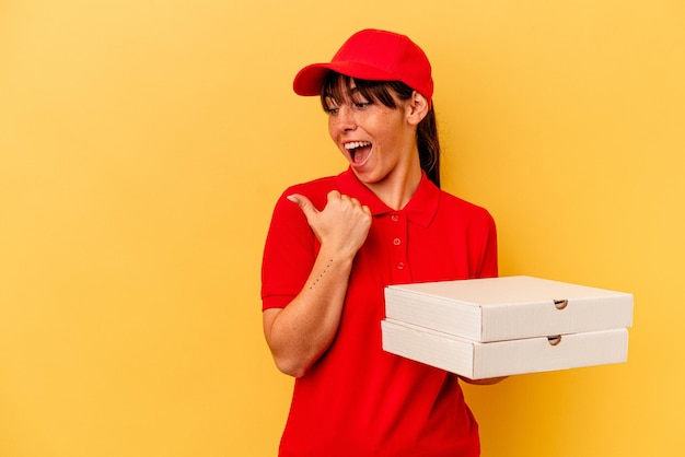 Mulher de entrega jovem segurando pizzas isoladas em pontos de fundo amarelo com o dedo polegar afastado, rindo e despreocupado.
