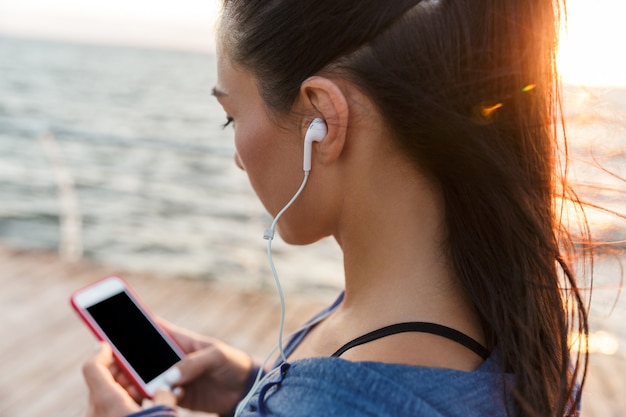 Foto mulher de esportes ao ar livre na praia, ouvindo música com fones de ouvido, usando o telefone celular.