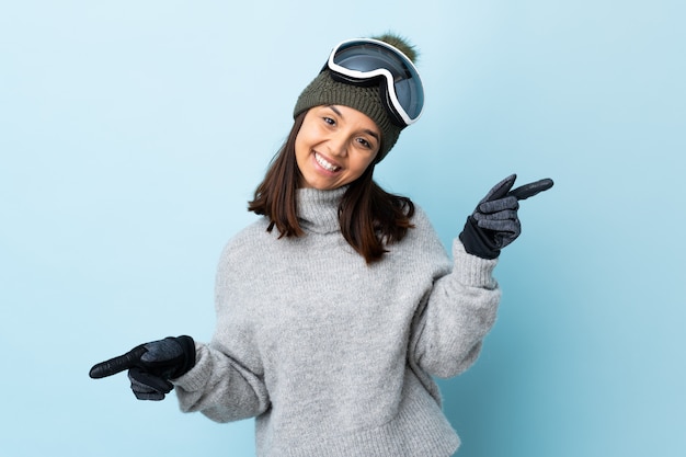 Mulher de esquiador de raça mista com óculos de snowboard sobre parede azul isolada, apontando o dedo para as laterais e feliz