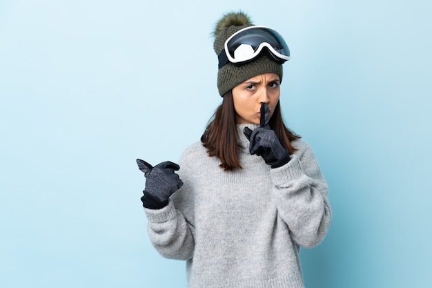 Mulher de esquiador de raça mista com óculos de snowboard sobre parede azul isolada, apontando para o lado e fazendo o gesto de silêncio