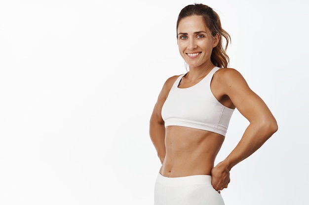 Mulher de fitness forte e apta com 40 anos de idade, abs e músculos sorrindo e parecendo confiante após o treino de fundo branco