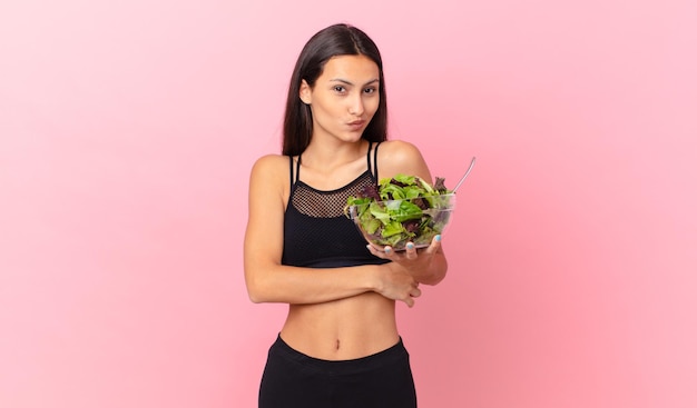 Mulher de fitness hispânica encolhendo os ombros sentindo-se confusa e incerta e segurando uma salada