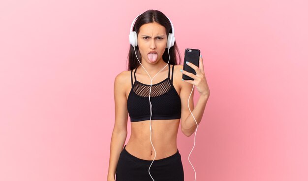 Mulher de fitness hispânica se sentindo enojada e irritada e língua de fora com fones de ouvido e um telefone