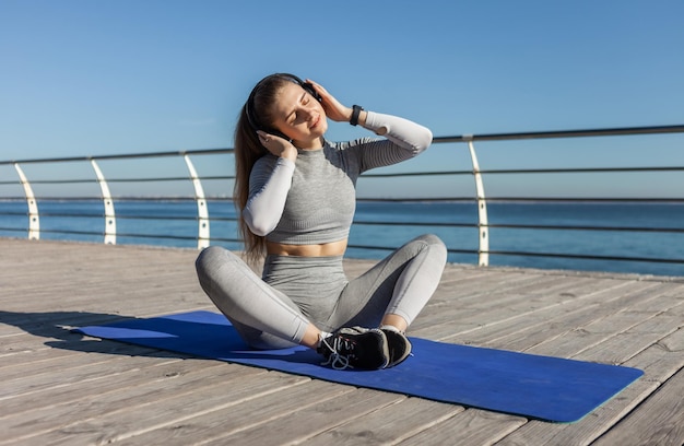 Mulher de fitness relaxada ouvindo música com fones de ouvido enquanto está sentado em um tapete na praia Esportes e relaxamento
