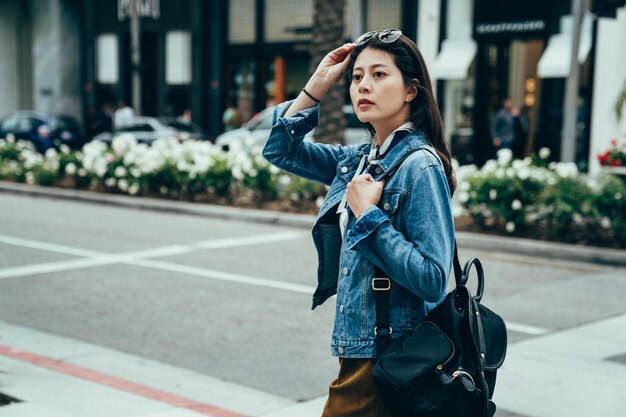 mulher de fundo desfocado carregando mochila está procurando sinais de trânsito para encontrar seu caminho. elegante mulher asiática segurando óculos de sol está andando na rua, percebendo algo estranho.