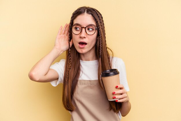 Mulher de garçom de restaurante segurando um café para viagem isolado em fundo amarelo tentando ouvir uma fofoca.