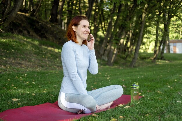 Mulher de gengibre atlética em roupas esportivas usando telefone celular no parque. Saúde, bem-estar, estilo de vida fitness.