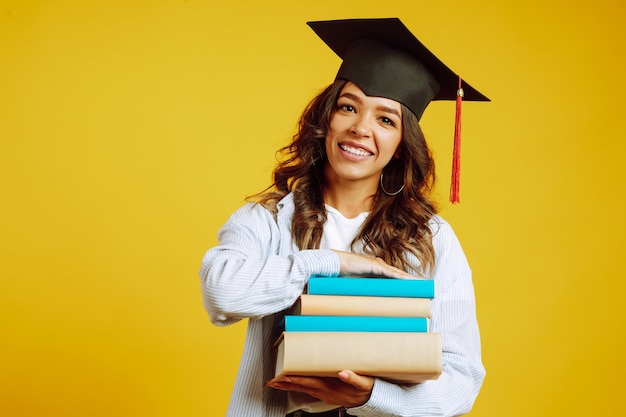 Foto mulher de graduação com um chapéu de formatura na cabeça, com livros em amarelo.