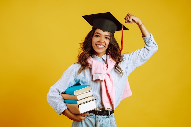 Mulher de graduação com um chapéu de formatura na cabeça, com livros em amarelo.