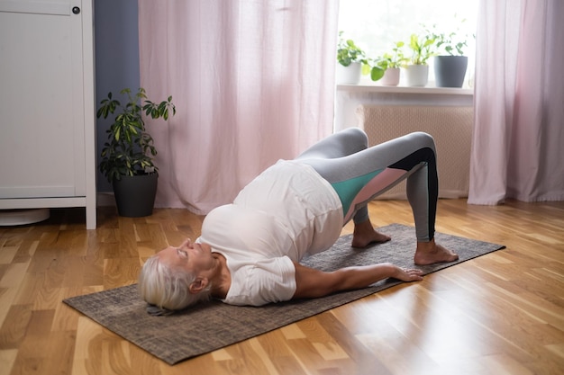 Mulher de ioga atlética sênior fazendo roda ardha chakrasana no tapete de ioga em casa