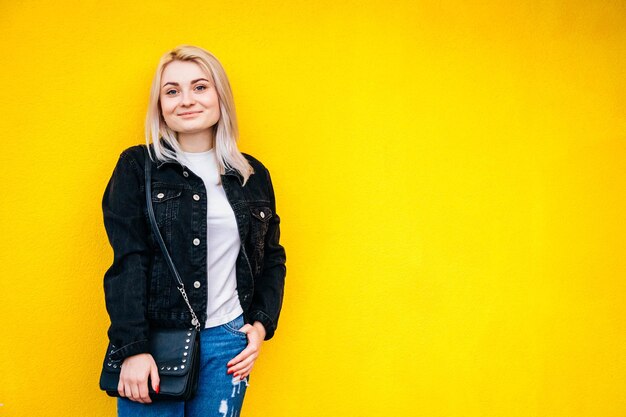 Foto mulher de jaqueta preta sorrindo, posando contra um fundo de parede amarela