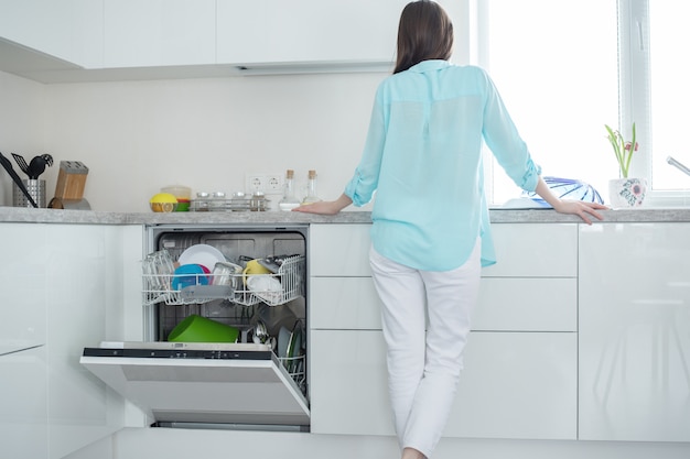 mulher de jeans branco e camisa fica de costas ao lado de uma máquina de lavar louça aberta no interior da cozinha