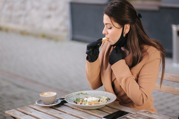 Mulher de luvas pretas segura talheres. Conceito do Quarantine Cafe. Alimentos ao ar livre em luvas de proteção.