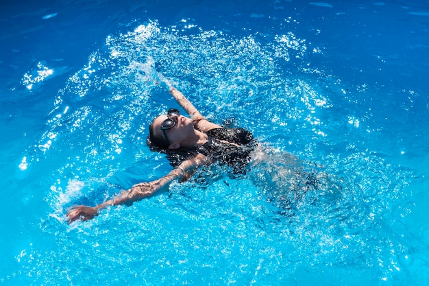 Mulher de maiô preto e óculos de sol nada de costas em águas claras na piscina Mulher bonita em uma piscina