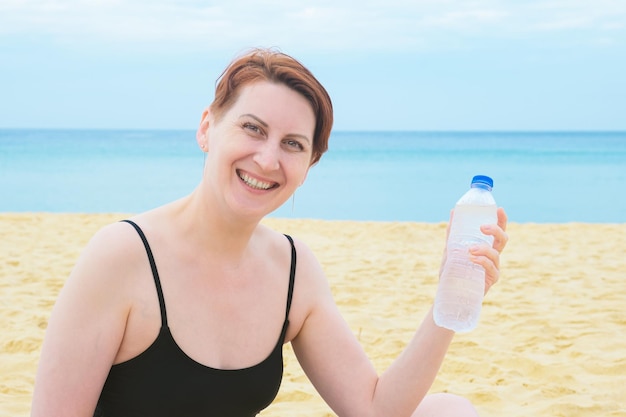 Mulher de maiô senta-se na praia e segura uma garrafa plástica de água potável na mão