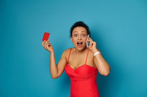 Mulher de maiô vermelho falando em um telefone celular e olhando para um cartão de plástico vermelho sobre fundo azul