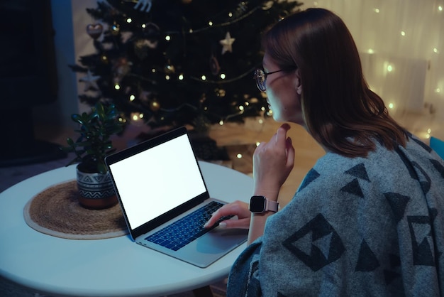 Mulher de maquete de laptop de tela branca em branco usando laptop e relógio inteligente em casa no fundo da árvore de natal e luzes