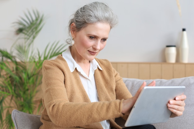 Mulher de meia idade alegre sentada no sofá, usando aplicativos de tablet de computador, olhando para a tela, lendo boas notícias na rede social, fazendo compras ou conversando online.