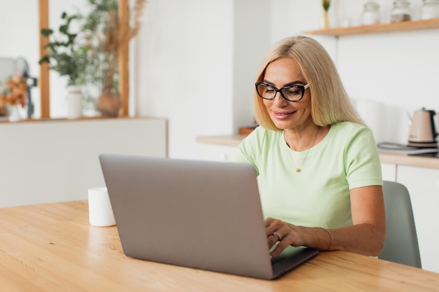 Mulher de meia idade atraente moderna tomando café e trabalhando no laptop na cozinha em casa