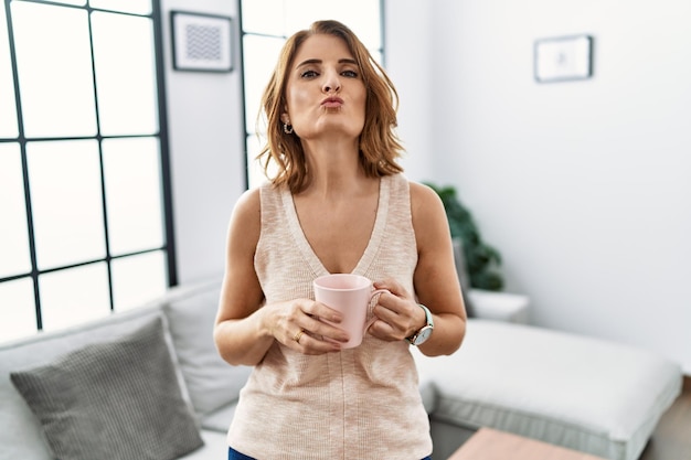Mulher de meia-idade bebendo uma xícara de café em casa olhando para a câmera mandando um beijo no ar sendo uma expressão de amor adorável e sexy
