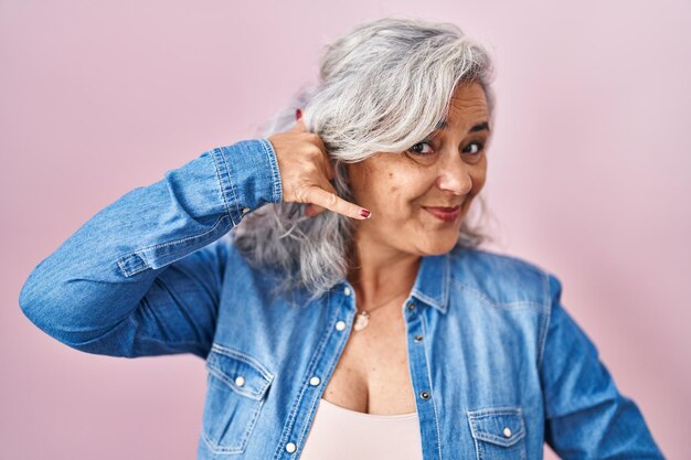 Foto mulher de meia-idade com cabelos grisalhos em pé sobre fundo rosa sorrindo fazendo gesto de telefone com a mão e os dedos como falar ao telefone comunicando conceitos