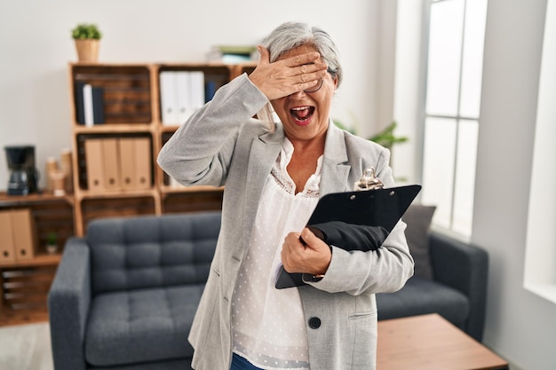 Mulher de meia-idade com cabelos grisalhos no escritório de consulta sorrindo e rindo com a mão no rosto cobrindo os olhos para o conceito cego de surpresa