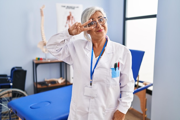 Mulher de meia idade com cabelos grisalhos trabalhando na clínica de recuperação de dor fazendo símbolo de paz com os dedos sobre o rosto, sorrindo alegre mostrando vitória